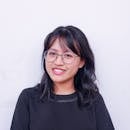 Portrait of a young woman with glasses and black hair smiling against a plain background.